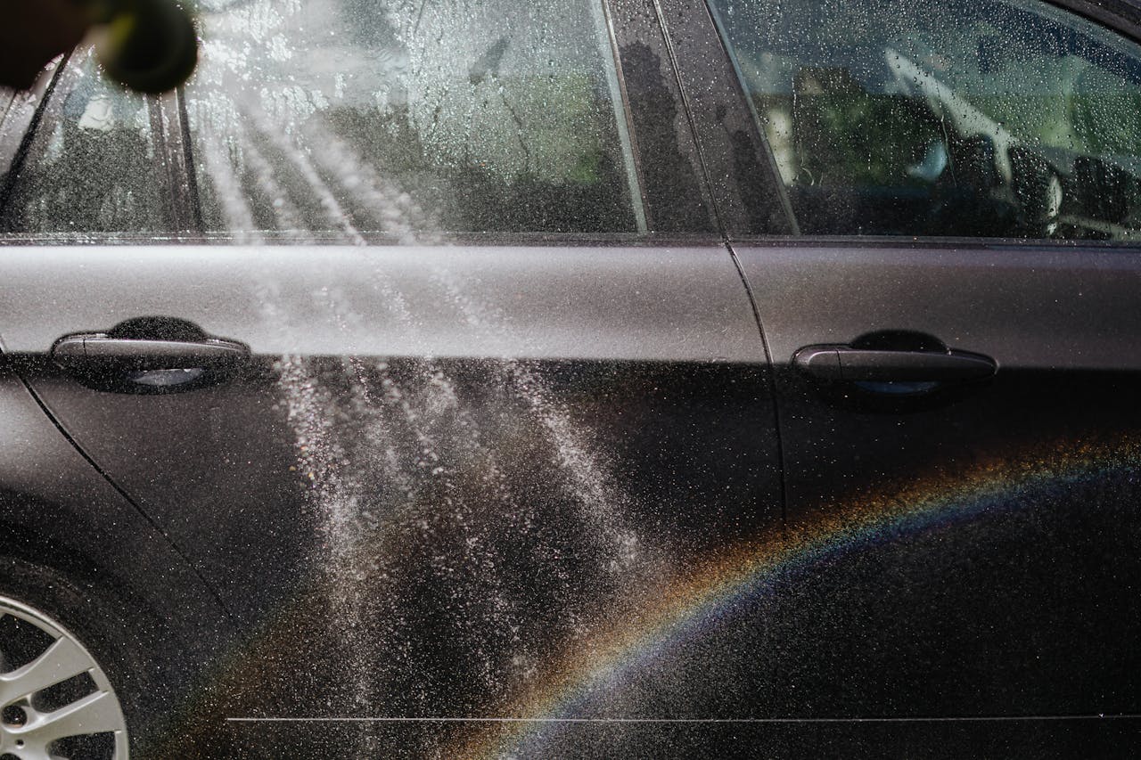 Close-up of a car being washed with a visible rainbow in sunlight.